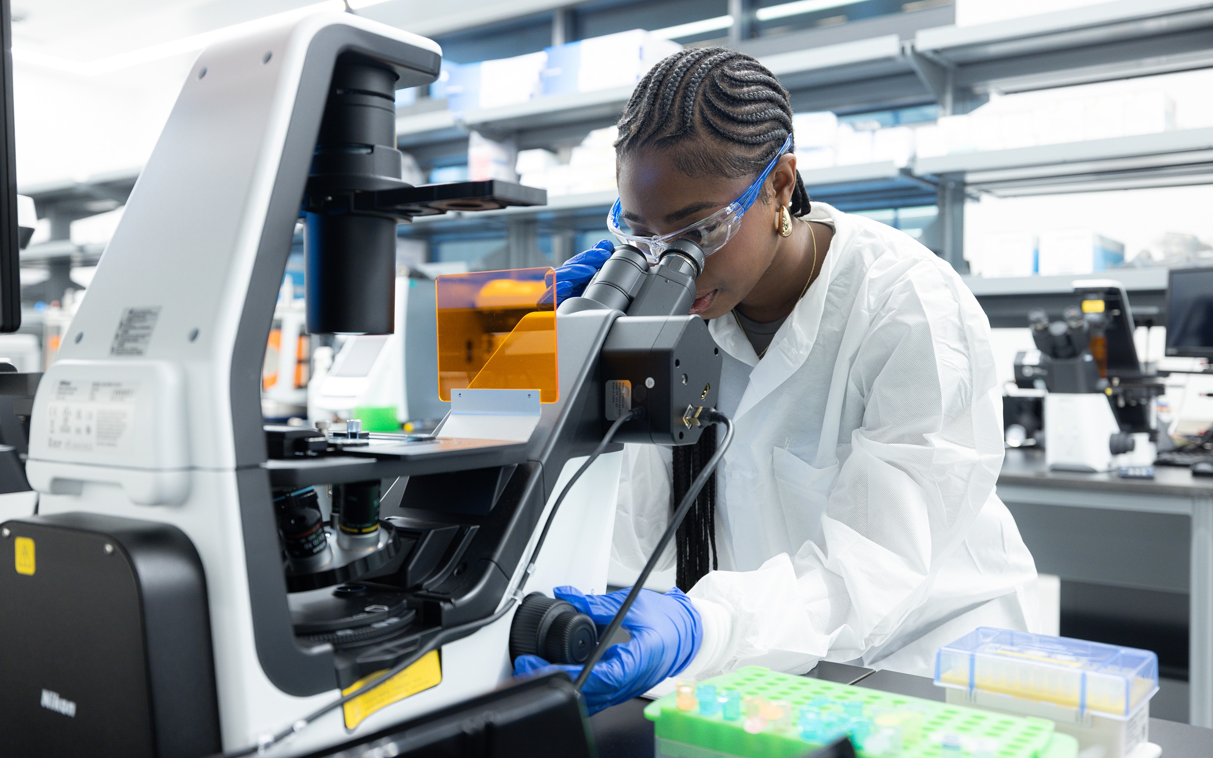 A BioTC trainee wearing safety glasses and gloves examines samples through a microscope.